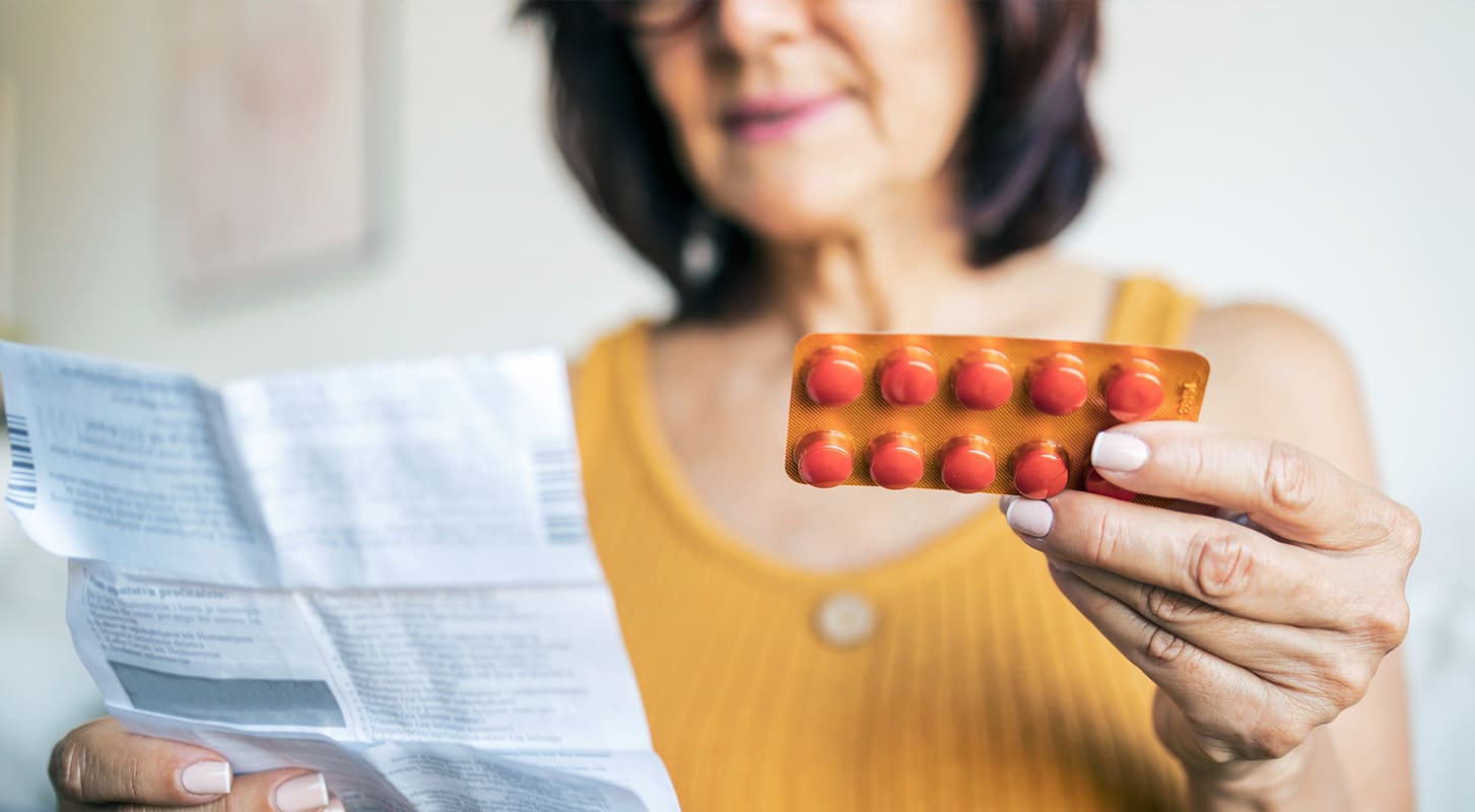 Close-up of a person holding a blister pack of orange pills in one hand and a medical prescription or drug information sheet in the other.